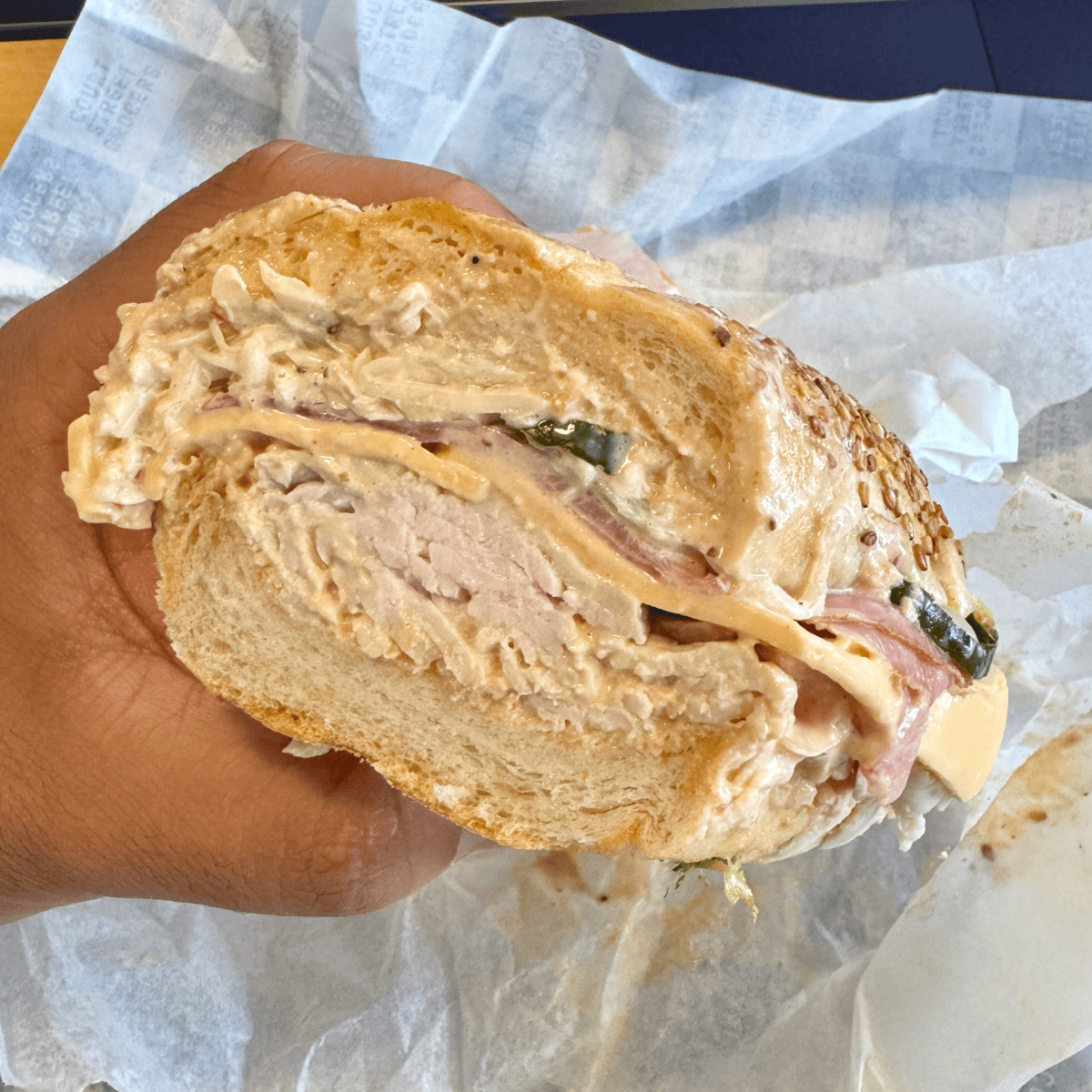 A close-up of a hand holding a large sandwich on a sesame seed roll, filled with sliced turkey, melted cheese, pickles, onions, and creamy sauce, with sandwich paper in the background.